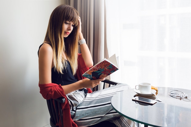 Indoor lifestyle portret van mooie brunette vrouw leesboek, zittend op een stoel en koffie drinken in zonnige lentemorgen. Ontbijt tijd.