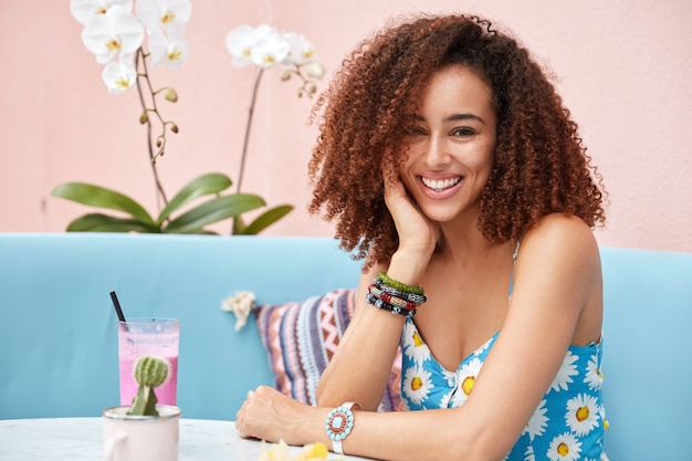 Indoor portret van Afro-Amerikaanse vrouw met borstelig krullend haar, zit aan tafel in cafetaria met cocktail, wordt in hoge geest, viert beginnende vakantie.