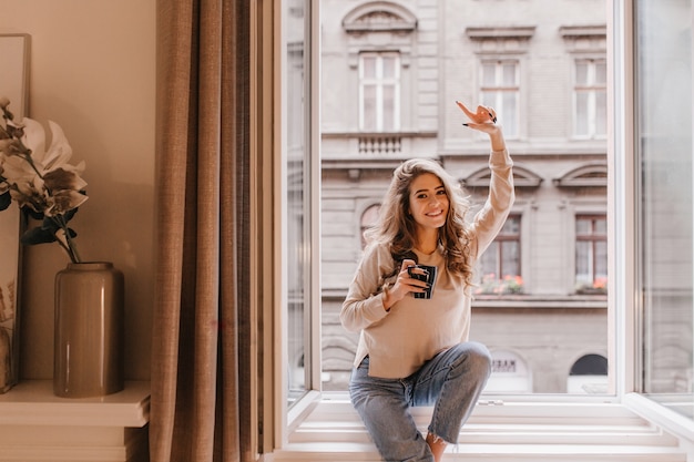 Indoor portret van bevallige blanke vrouw poseren met plezier op vensterbank