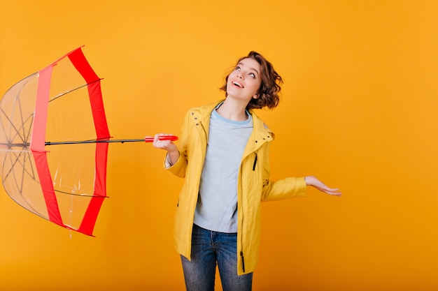 Indoor portret van charmant meisje opzoeken tijdens regenachtig weer. blanke bleke vrouw in gele jas poseren met parasol en blij gezichtsuitdrukking.