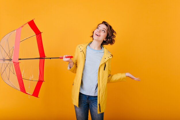 Indoor portret van charmant meisje opzoeken tijdens regenachtig weer. blanke bleke vrouw in gele jas poseren met parasol en blij gezichtsuitdrukking.