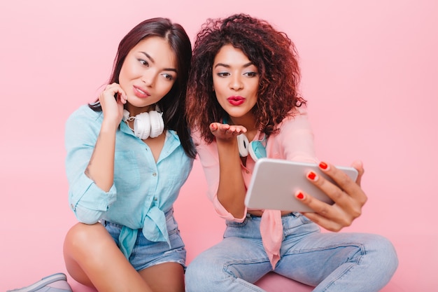 Indoor portret van charmante krullende vrouw met witte smartphone selfie maken in de buurt van vriend. Mooie brunette dames in zomerkleren foto van zichzelf nemen.