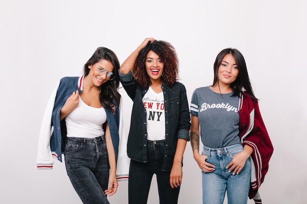 Indoor portret van drie opgewonden vrouwelijke studenten in trendy kleding met plezier samen na de lessen. Krullend meisje in denimkleding tijd doorbrengen met donkerbruine vrienden en lachen.