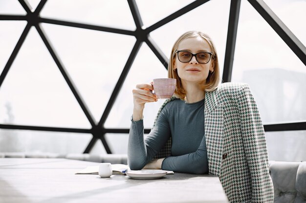 Indoor portret van een jonge zakenvrouw die in een café zit en een kopje koffie drinkt. Blond meisje met zonnebril en jas