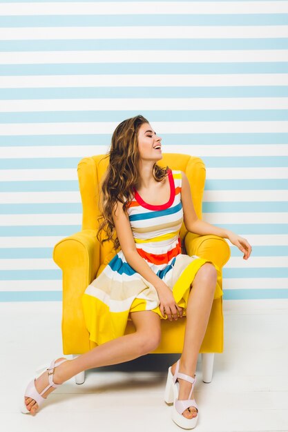 Indoor portret van geïnspireerde schattig meisje met hoge hakken en gestreepte kleurrijke jurk. Sierlijke jonge vrouw met gebruinde huid op gele fauteuil staan in haar kamer en lachen.