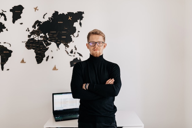 Indoor portret van knappe blonde man met bril en zwarte trui poseren over witte muur met wereldkaart en laptop op desktop.