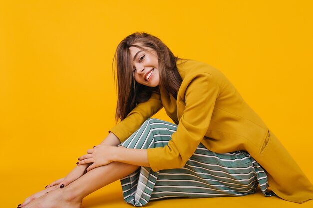 Indoor portret van lachen schattig meisje in stripted broek omarmen haar benen. Sierlijke blootsvoets vrouw poseren met plezier.