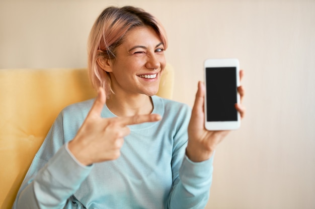 Indoor portret van mooie positieve jonge Europese vrouw zittend op een stoel met haar nieuwe slimme telefoon