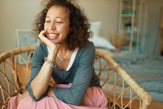 Indoor Portret van mooie verlegen jonge Spaanse vrouw met donkere huid en lang krullend haar ontspannen thuis, zittend in rotan stoel breed glimlachend