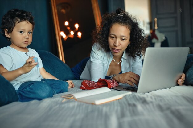 Indoor Portret van schattige gemengd ras kleine jongen zittend op bed en het maken van tekeningen terwijl zijn jonge moeder met behulp van draagbare computer voor extern werk.