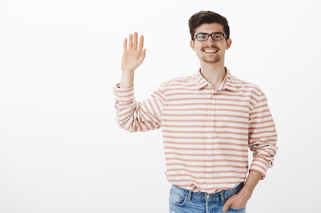 Gratis foto indoor portret van vriendelijke gewone europese man met baard en snor in nerdy bril, palm omhoog en zwaaien, hallo zeggen tegen teamleden, begroetend personeel