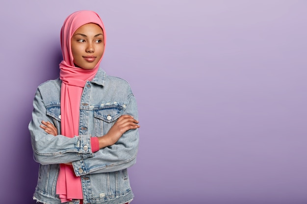 Indoor shot doordachte mooie Arabische vrouw staat binnen met gekruiste handen, gefocust weg, overweegt toekomstige plannen, draagt hijab en denim jas, geïsoleerd over paarse muur.