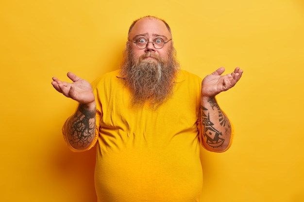 Gratis foto indoor shot van aarzelende bebaarde man met overgewicht haalt zijn schouders op en staat onbewust, heeft dikke baard, grote bierbuik, gekleed in geel t-shirt, ronde bril, gezichten moeilijke keuze.