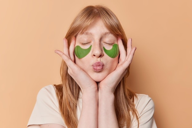 Indoor shot van blij roodharig meisje raakt gezicht zachtjes sluit ogen houdt lippen afgerond past groene hydrogel patches onder ogen voor hydratatie geïsoleerd over bruine achtergrond Beauty concept