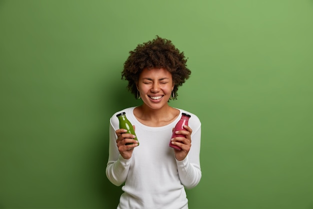 Indoor shot van erg blij lachende vrouw houdt flessen gemengde spinazie en aardbei smoothie, geniet van detox drank drinken, poses met vers bereide verse drank, geïsoleerd op groene achtergrond