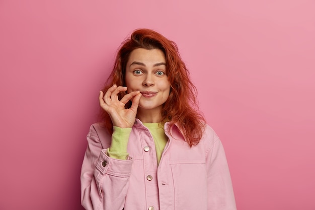 Indoor shot van geheime roodharige jonge vrouw ritst haar mond dicht, belooft geheim te houden, roddelt veilig, houdt vingers bij de wang