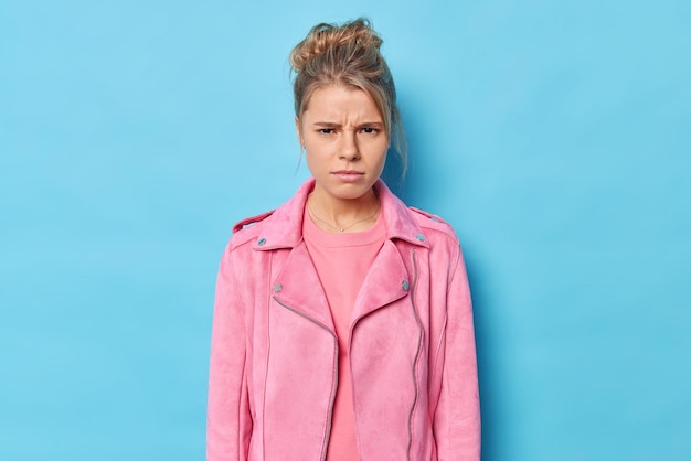 Indoor shot van ontevreden jonge europese vrouw fronst gezicht met woede heeft mokkende sombere uitdrukking draagt roze jas ziet er beledigd of beledigd uit geïsoleerd over blauwe achtergrond wacht op verontschuldiging