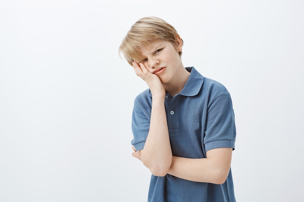 Indoor shot van ontevreden ongelukkige jonge jongen met blond haar in blauw t-shirt, gezicht palm en grimassen maken, geïrriteerd of beu zijn met huiswerk