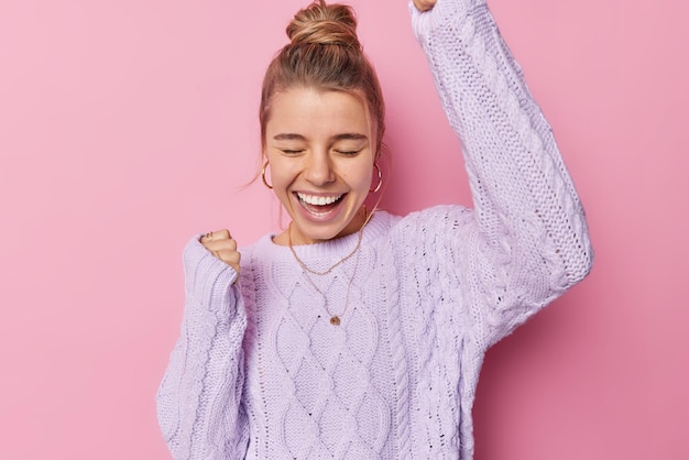 Gratis foto indoor shot van optimistische jonge vrouw met knot hairstye maakt ja gebaar lacht positief viert prestaties draagt gebreide paarse trui geïsoleerd over roze achtergrond eindelijk succes