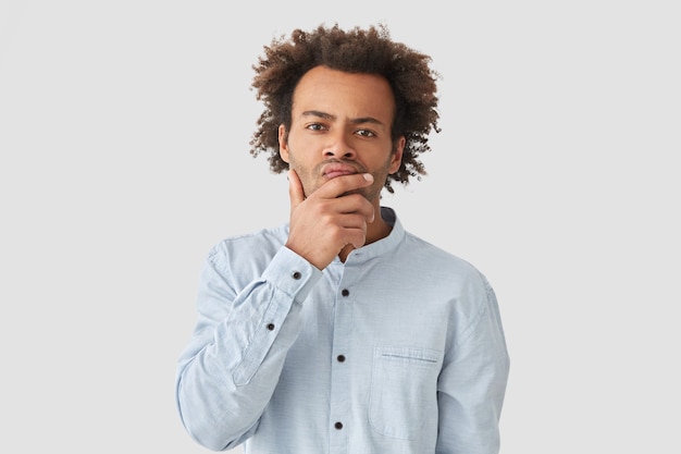 Indoor shot van peinzende Afro-Amerikaanse man houdt kin en kijkt met een ernstige uitdrukking, denkt over zijn probleem, draagt een elegant-shirt, heeft een Afro-krullend kapsel, geïsoleerd