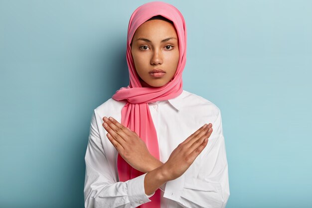 Indoor shot van serieuze moslimvrouw maakt ontkenning handgebaar, houdt armen gekruist over de borst, toont stopbord, draagt hoofddoek, volgt religieuze dresscode, geïsoleerd over blauwe muur