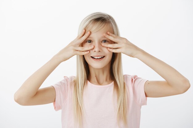 Indoor shot van verbaasd verrast schattige dochter met brede, gelukkige glimlach, vingers bij de ogen vasthoudend en grijnzend van positieve scène, vrolijk zijn, groot feest bijwonen ter ere van haar over grijze muur