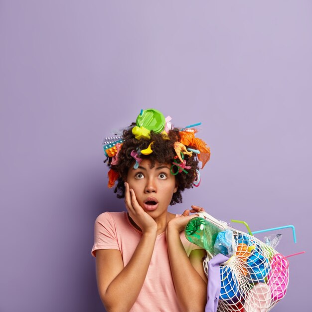 Indoor shot van verbaasde donkere vrouw kijkt met verlegenheid boven, raakt wang, heeft vuilnis in het haar houdt netzak vol afval draagt casual t-shirt voelt zich teleurgesteld. Ecologie, vervuiling