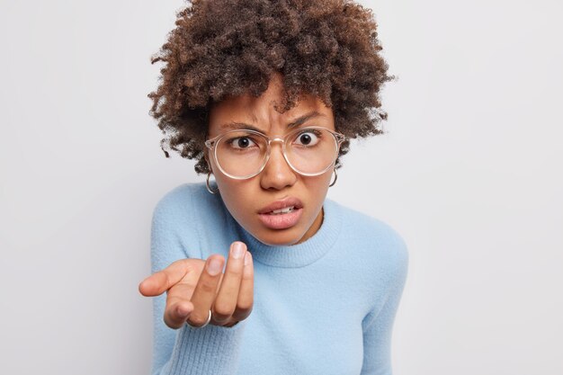 Indoor shot van verontwaardigd verbaasd krullend haired Afro-Amerikaanse vrouw shugs schouder heeft aarzelende uitdrukking draagt een bril casual coltrui geïsoleerd op witte achtergrond. Dus wat zei je.