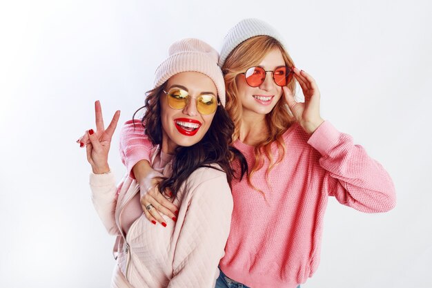 Indoor studio afbeelding van twee meisjes, gelukkige vrienden in stijlvolle roze kleding en hoed spelling grappig samen. Witte achtergrond. Trendy hoed en bril die vrede tonen.