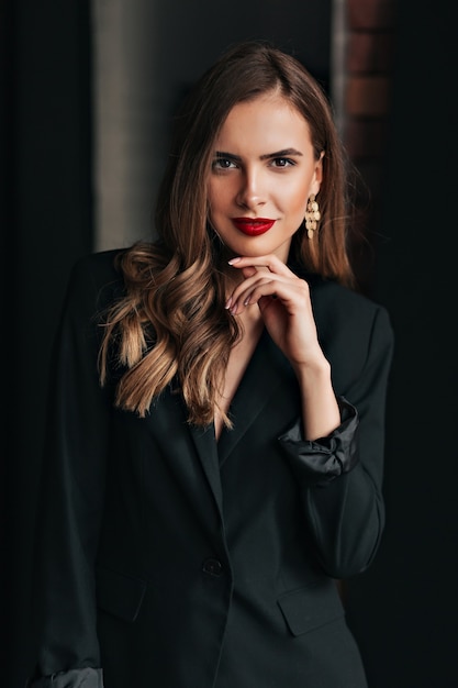Indoor studio-opname van aantrekkelijke mooie vrouw met lichtbruin haar, gekleed in zwarte jas met rode lippen