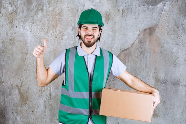 Ingenieur in gele uitrusting en helm die een kartonnen doos vasthoudt en een positief handteken toont