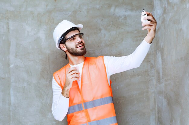 Ingenieur man in witte helm en beschermende bril met een wekker en een kopje drank.