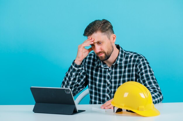 Ingenieur man met hoofdpijn houdt hand op voorhoofd door voor atblet op blauwe achtergrond te zitten