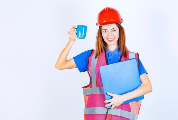 Ingenieursvrouw in rode helm die een blauwe omslag houdt en een kop van drank heeft.