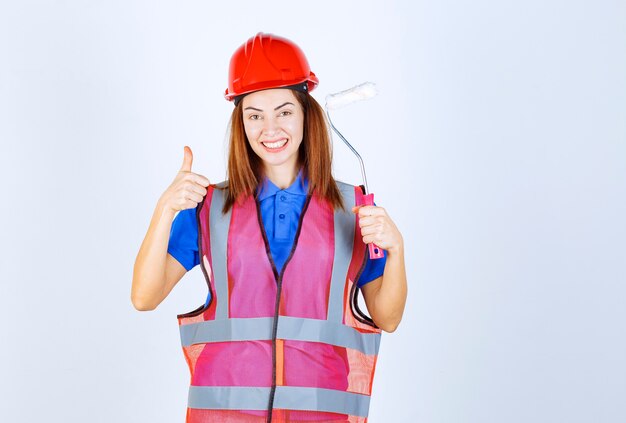 Ingenieursvrouw in uniform die een witgekleurde verfroller vasthoudt en een tevredenheidsteken toont.