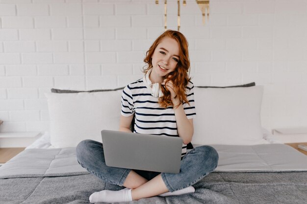 Innemend kaukasisch meisje in spijkerbroek die laptop gebruikt terwijl ze op bed zit Prachtige gemberdame in koptelefoon die geniet van weekend