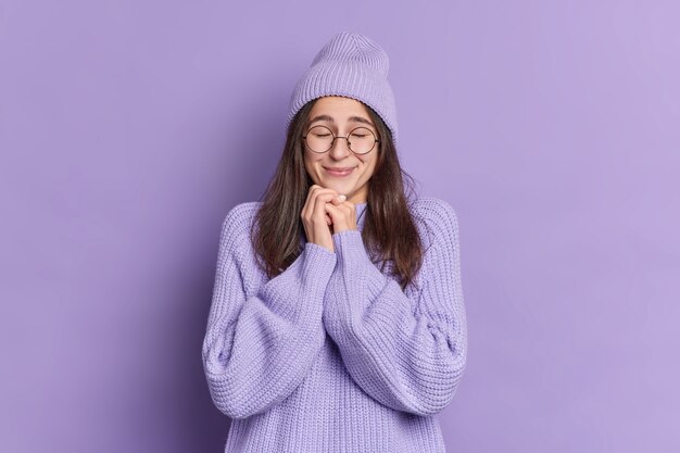 Inschrijving mooie donkerharige jonge vrouw met Europese uitstraling houdt de handen bij elkaar in de buurt van het gezicht sluit de ogen blij om hartverwarmende woorden te horen gekleed in gebreide kleding.