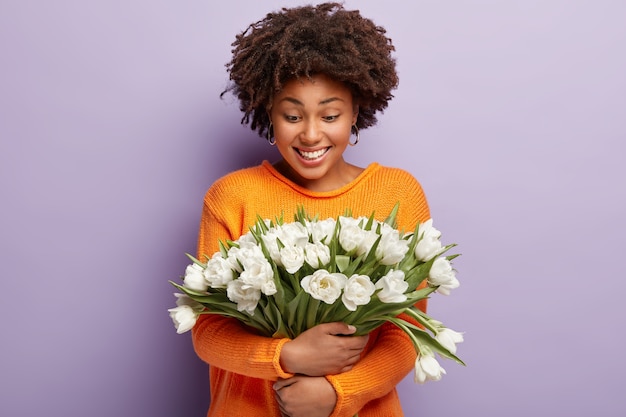 Inschrijving positieve Afro-Amerikaanse dame omhelst teder witte bloemen, lacht zachtjes en kijkt naar tulpen, draagt oranje trui