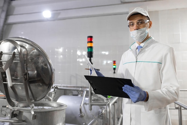 Inspecteur met een masker en een scrub staat met een foldertablet in zijn handen bij de zuivelfabriek