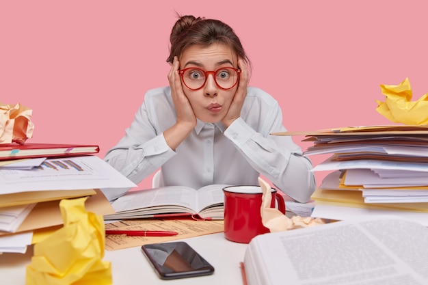 Gratis foto intelligente student kijkt aandachtig recht in de camera, raakt de wang met beide handpalmen aan, draagt een wit overhemd, is bezig met het schrijven van cursuspapier, schoolboeken, papieren op de werkplek