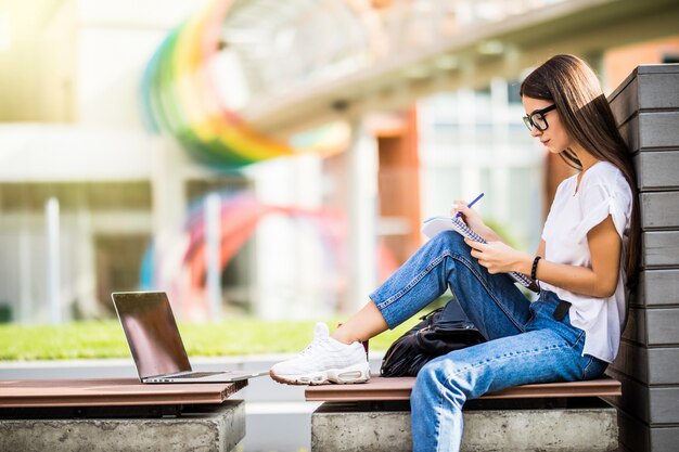 Intelligente vrouw die in glazen nota's in planner maken en laptop doorbladeren terwijl het zitten op bank in werf van modern gebouw in stad