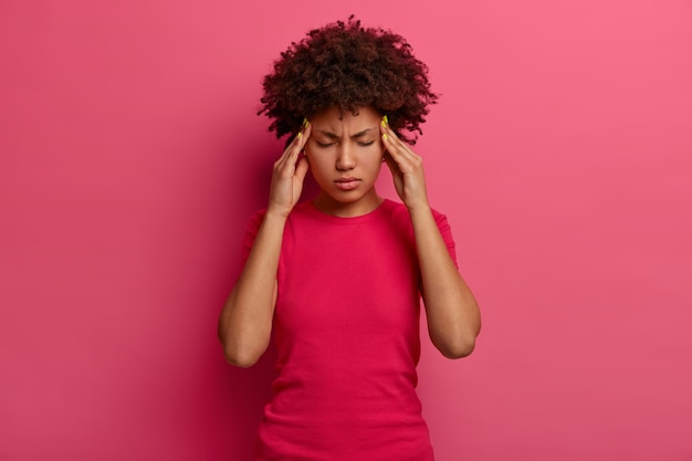 Gratis foto intens geperste duizendjarige vrouw met een donkere huid voelt zich moe en duizelig, houdt de handen op de slapen, lijdt aan ondraaglijke hoofdpijn, kan niet tegen pijn, grimassen van migraine, geïsoleerd op een roze muur