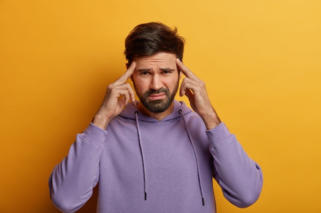 Intens ongeschoren man probeert zich te concentreren en iets te herinneren, houdt wijsvingers op de slapen, lijdt aan migraine, heeft een ontevreden uitdrukking, draagt een casual sweatshirt, geïsoleerd op een gele muur