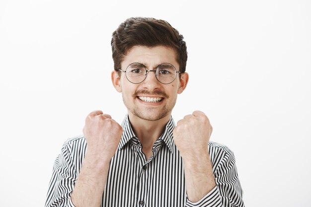 Ja, we hebben het project op tijd afgerond. Portret van knap volwassen mannelijk model in brillen en gestreept overhemd, gebalde vuisten opheffen, zelfverzekerd zijn, succes en overwinning vieren over grijze muur