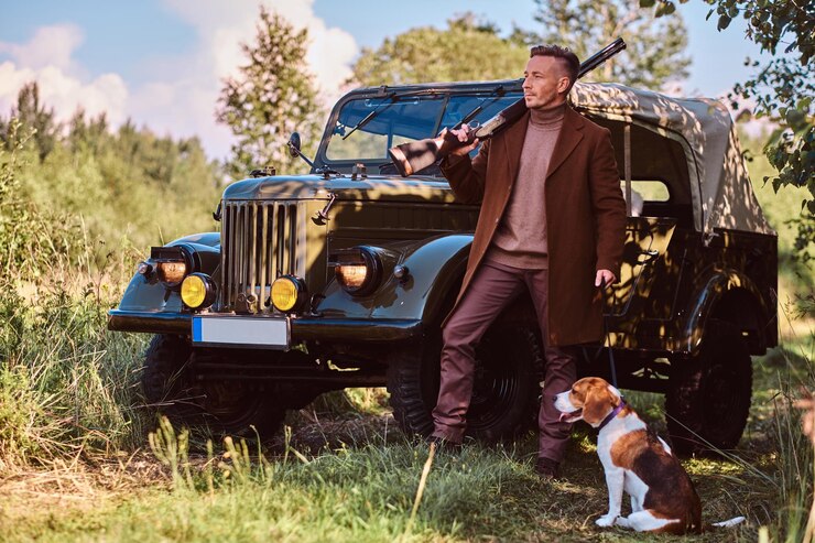Gratis Foto jager in elegante kleding houdt een jachtgeweer vast en staat samen met zijn beagle-hond in de buurt van een retro militaire auto in het bos.