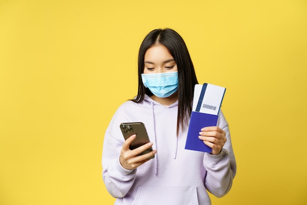 Japans meisje met gezichtsmasker, met haar paspoort en vliegtickets, smartphone-app gebruikend, op vakantie gaan, reizen tijdens covid pandemie, gele achtergrond