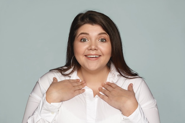 Je bedoelt mij. gefascineerde verbaasde jonge plus-size vrouw in wit overhemd wijzend naar zichzelf, hand in hand op haar borst en breed glimlachend als gekozen voor baanpositief onder andere kandidaten