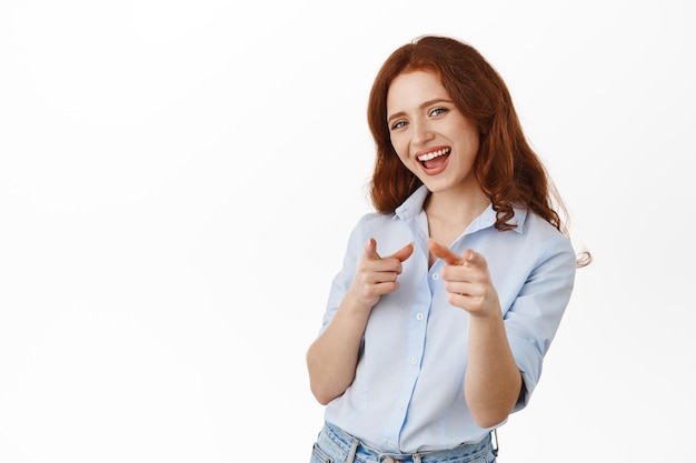 Je hebt dit, gefeliciteerd. Glimlachende roodharige vrouw die met de vingers naar de camera wijst, je uitnodigt, persoon prijst, nomineert, staande tegen een witte achtergrond