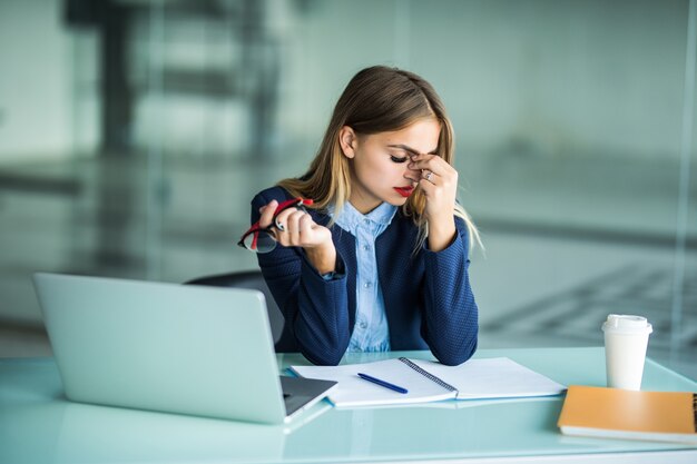 Je moe en gestrest voelen. Gefrustreerde jonge vrouw die de ogen gesloten houdt en de neus masseert terwijl ze op haar werkplek op kantoor zit