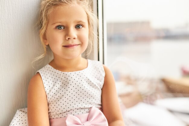 Jeugd en onschuldig concept. Portret van charmante schattig klein meisje met verzamelde blonde haren en grote mooie ogen zitten bij raam met gelukkige gezichtsuitdrukking en glimlachen